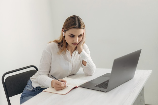 Una giovane bella ragazza in cuffia impara con la chat su internet un insegnante skype si prepara per un esame una studentessa impara online orologi un webinar prende appunti guarda un laptop apprendimento a distanza