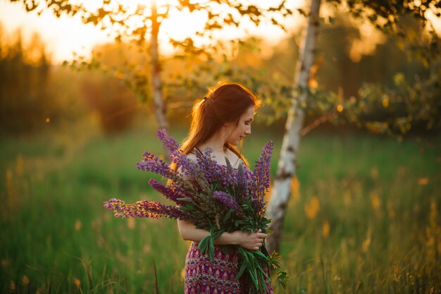 Young beautiful girl holding a large flower with purple lupine in a flowering field. Blooming lupine flowers.