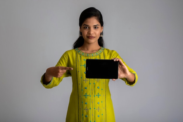 Young beautiful girl holding and showing blank screen of smartphone or mobile or tablet phone on a gray wall.