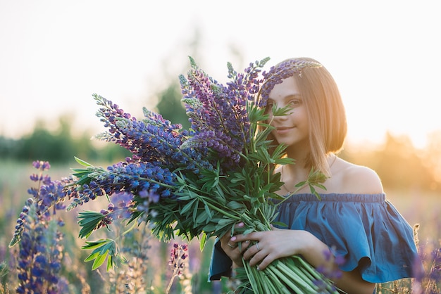 美しい少女は、開花フィールドで紫色のルピナスの大きな花束を保持しています。夏の写真。日没時の写真