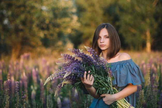 Young beautiful girl holds a large bouquet of purple lupins in a flowering field. Summer photo. Photo at sunset