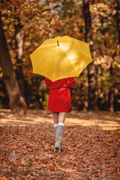 Фото Молодая красивая девушка в красном платье идет одна под желтым зонтиком в парке золотой осенью. вид сзади.