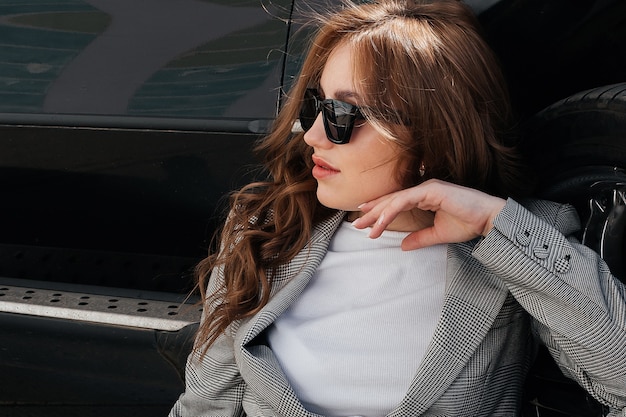 A young, beautiful girl is sitting on the asphalt near her black car in a suit and glasses.