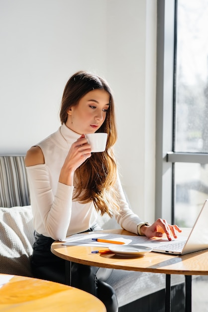 若い美しい女の子がカフェに座って、コンピューターで働いて、ソーシャルネットワークで通信しています。