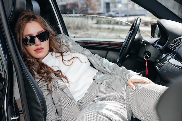 A young, beautiful girl is sitting in the front seat of a car. A stylish girl in a suit and glasses in a black leather car salon