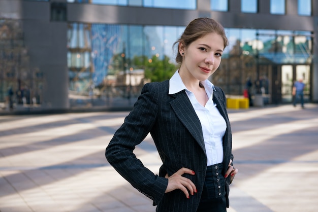 A young beautiful girl in a jacket and white shirt poses against the background of an office building on a sunny day