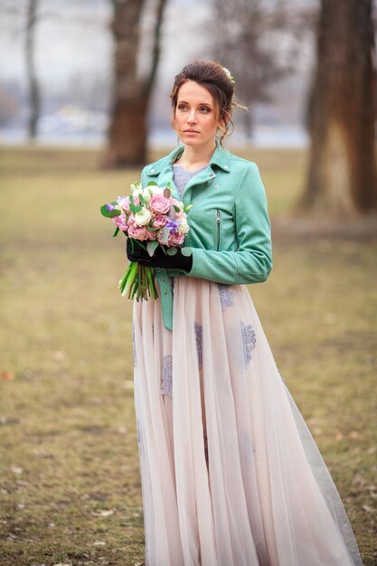 Young beautiful girl in a long dress and a green jacket and with a bouquet of flowers in her hands walks among the trees in the park. She is happy and smiling.