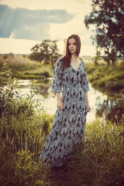 Young beautiful girl in a long dress standing against the sky and nature during the sunset