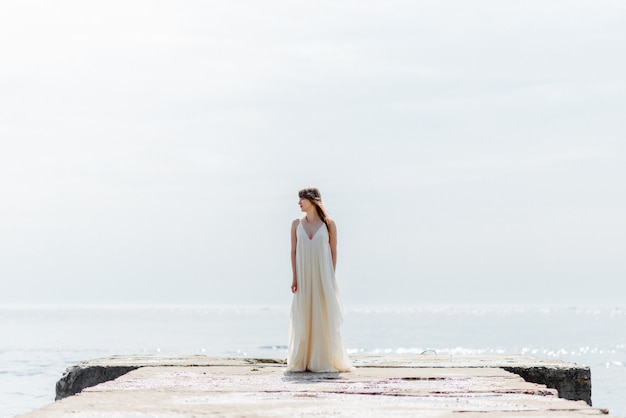 Una giovane e bella ragazza in un lungo abito bianco cammina lungo la spiaggia e il molo contro il mare.