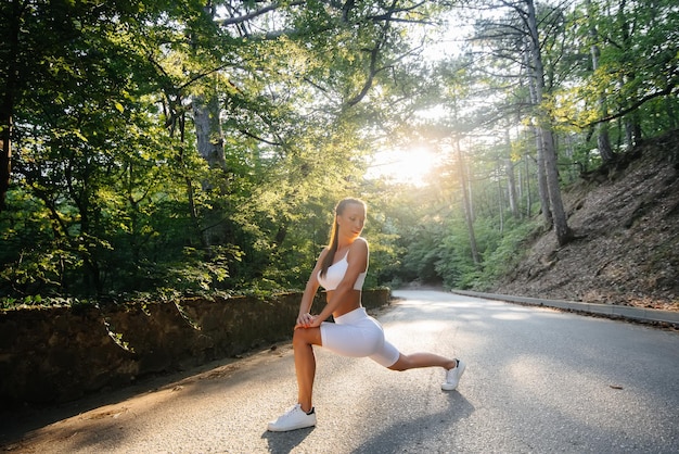 A young beautiful girl makes lunges and warm-up before running\
training, on the road in a dense forest, during sunset. a healthy\
lifestyle and running in the fresh air.