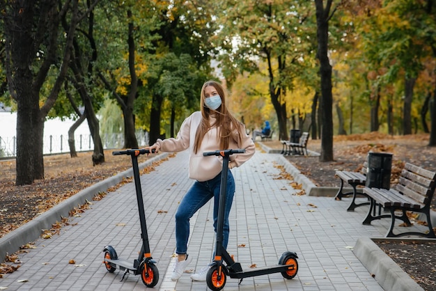 暖かい秋の日に、マスクをした若い美しい女の子が電動スクーターで公園に乗っています。公園で散歩する。