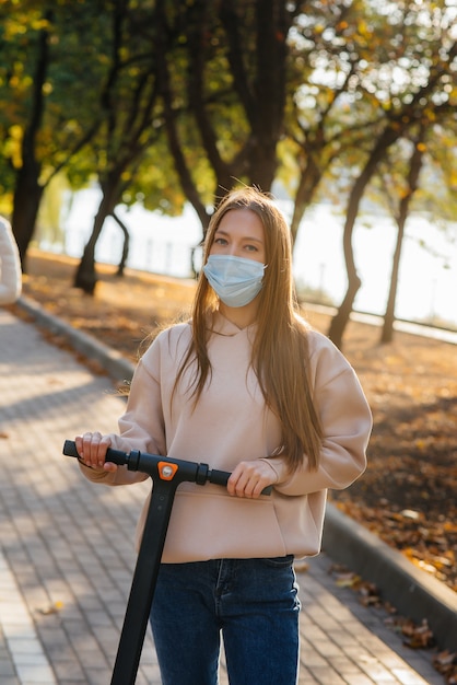 Una giovane bella ragazza con una maschera sta cavalcando nel parco su uno scooter elettrico in una calda giornata autunnale. camminare nel parco.