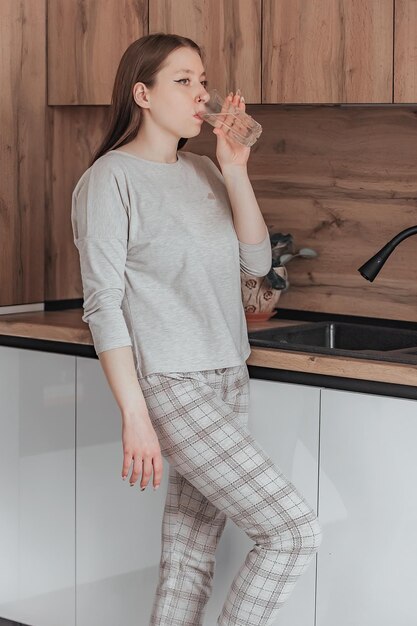 A young beautiful girl in pajamas drinks clean water in the kitchen in the morning