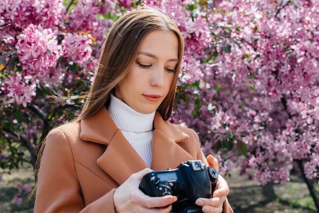 Молодая красивая девушка-фотограф ходит и фотографирует на фоне цветущей яблони. Хобби, отдых.