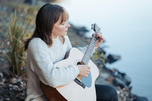 사진 강 근처에서 기타를 연주하는 아름다운 젊은 소녀 강을 바라보며 기타를 연습하는 소녀