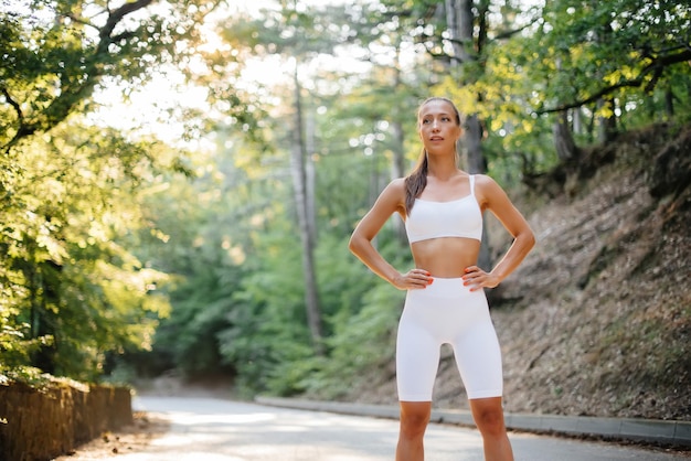 A young beautiful girl poses before running training, on the road in a dense forest, during sunset. A healthy lifestyle and running in the fresh air.