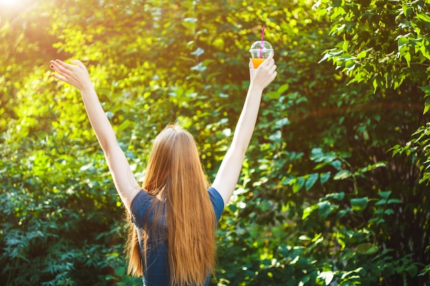 Young beautiful girl pulls her hands to the sun, holding in her hand juice.