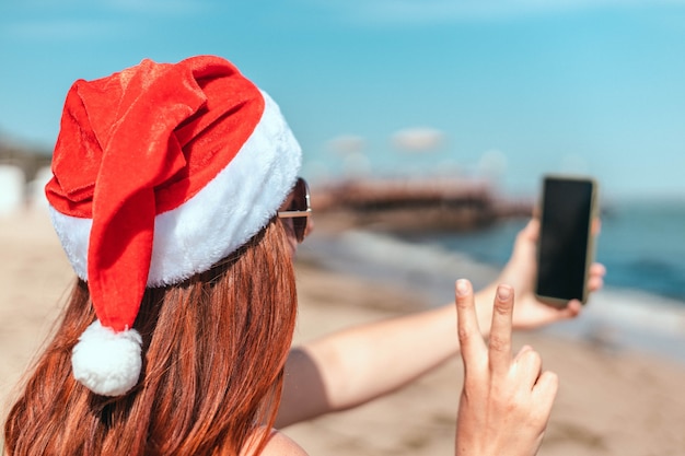 Young beautiful girl in a red santa claus hat and swimsuit makes a selfie on the seashore