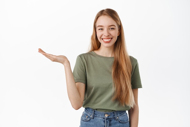 La giovane bella ragazza che mostra all'elemento che alza la mano aperta come se tenesse qualcosa di esposizione pubblicizza il prodotto su fondo bianco che si leva in piedi contro il fondo bianco