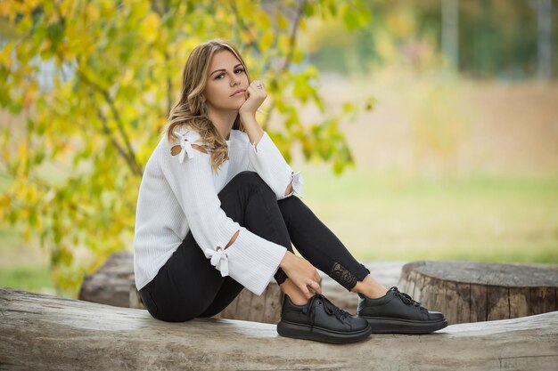 A young beautiful girl sits on a log in an autumn park against the background of yellow foliage