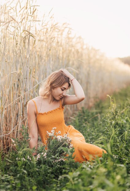 A young beautiful girl sits on a wheat field and holds a bouquet of daisies in her hands.