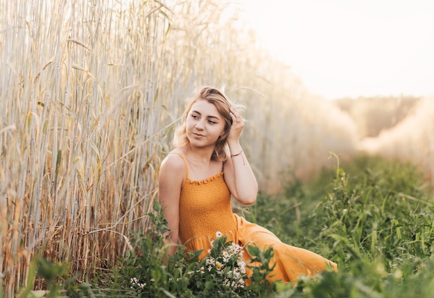 A young beautiful girl sits on a wheat field and holds a bouquet of daisies in her hands.