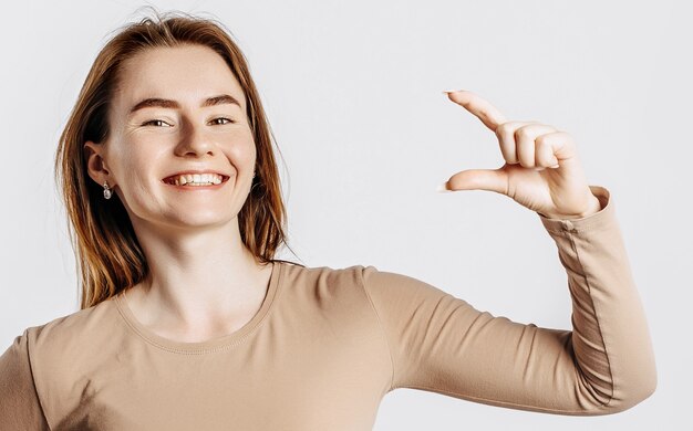 Young beautiful girl smiles and shows small size measure with her hand gesture on a white isolated background. A woman points to an idea, a place for advertising. Positive brunette in a beige jumper.