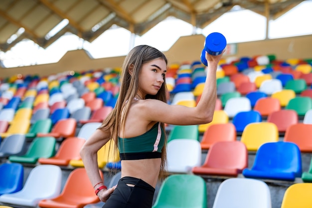 夏に市のスタジアムでダンベルを使って運動するスポーツウェアの若い美しい少女。スポーツコンセプト