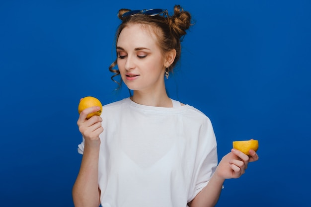 Молодая красивая девушка стоит на синем фоне, держа в руке лимоны и кусая