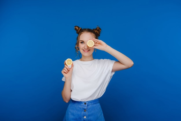 A young beautiful girl standing on a blue background holds lemons in her hands and covers her eyes with them