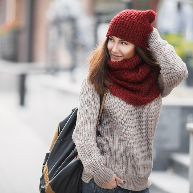 молодая красивая девушка-студентка в вязаном свитере, красном шарфе и шляпе и рюкзаке