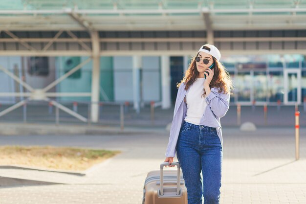 Young beautiful girl talking on the phone near a big bus stop with a suitcase