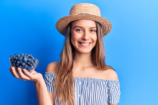 Young beautiful girl wearing summer hat holding bowl with blueberries looking positive and happy standing and smiling with a confident smile showing teeth