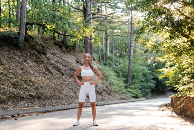 A young beautiful girl in white clothes poses before a running\
workout on a road in a dense forest during sunset healthy lifestyle\
and running in the fresh air youth