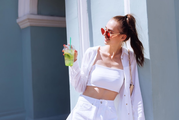 Young beautiful girl in white clothes and red glasses drinks a summer refreshing cocktail