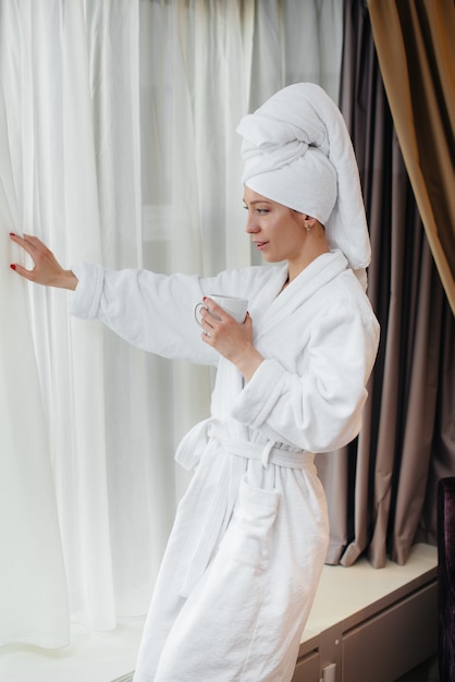 A young beautiful girl in a white coat drinks coffee in her hotel room.