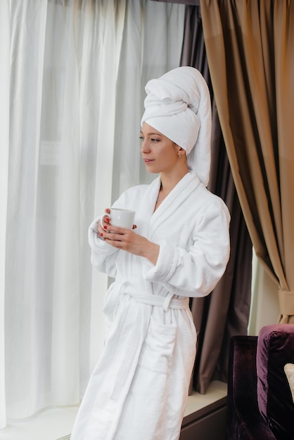 A young beautiful girl in a white coat drinks coffee in her hotel room.