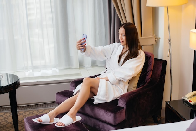 A young beautiful girl in a white coat takes a selfie on her phone in her hotel room.