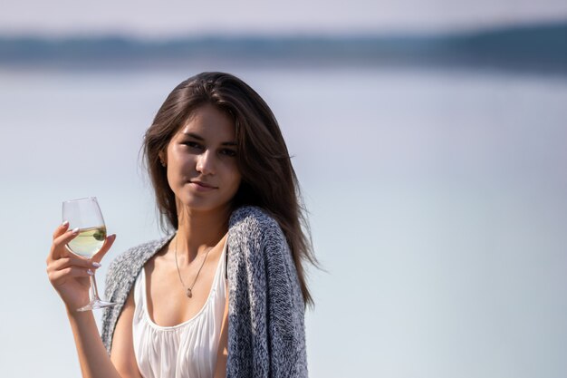 Young beautiful girl in white dress drinks wine from a glass. She is on the shore of the lake and enjoy a picnic and outdoor recreation.