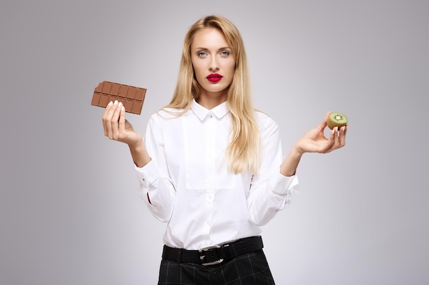 Giovane bella ragazza in camicia bianca tiene cioccolato e kiwi ritratto isolato in studio concetto di cibo sano e spazzatura