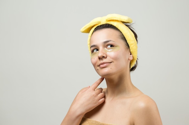 La giovane bella ragazza in un asciugamano bianco sulla testa indossa cerotti in gel di collagene sotto gli occhi maschera sotto il viso per il trattamento degli occhi