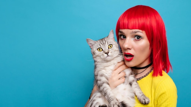 A young beautiful girl with a cat in her arms isolated on a blue background Expressive and funny expression on the face The friendship of the pet and the owner