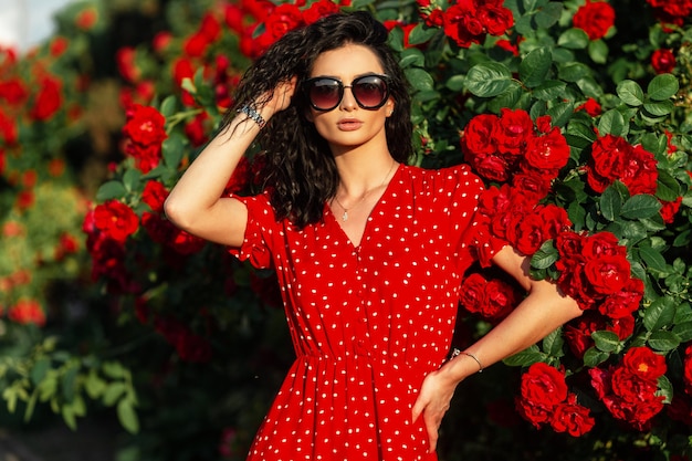 Young beautiful girl with fashionable sunglasses in a red dress with polka dots stands near a bush with rose flowers