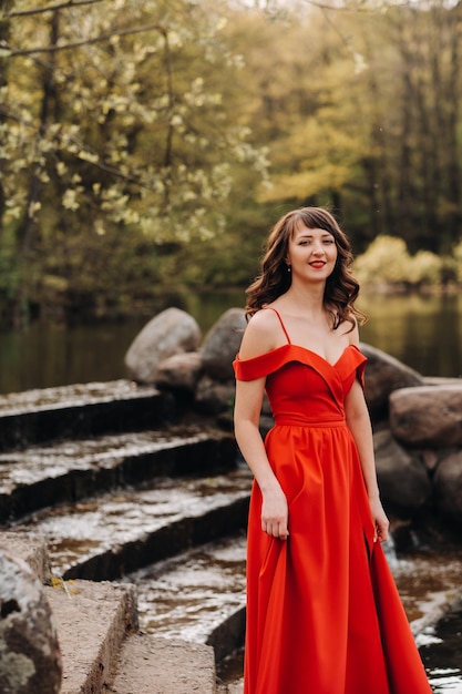 Молодая красивая девушка с длинными каштановыми волосами, в длинном красном платье спускается по лестнице у озера,