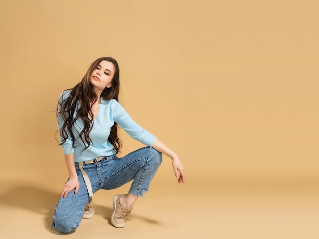 Young beautiful girl with long curly hair in a blue sweatshirt and blue jeans stands on one knee on a pastel orange.