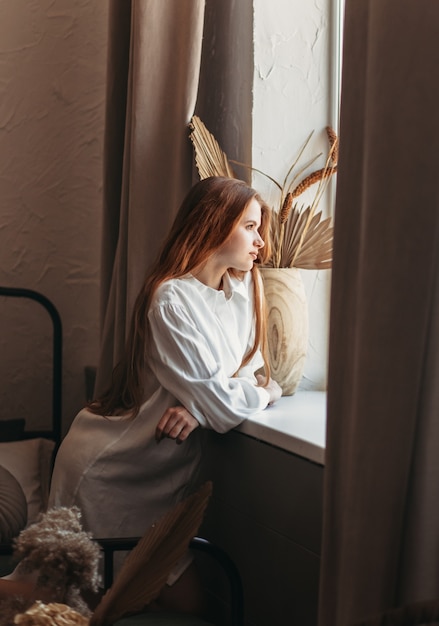 Young beautiful girl with long hair looks out the window on the bed. Vertical photo