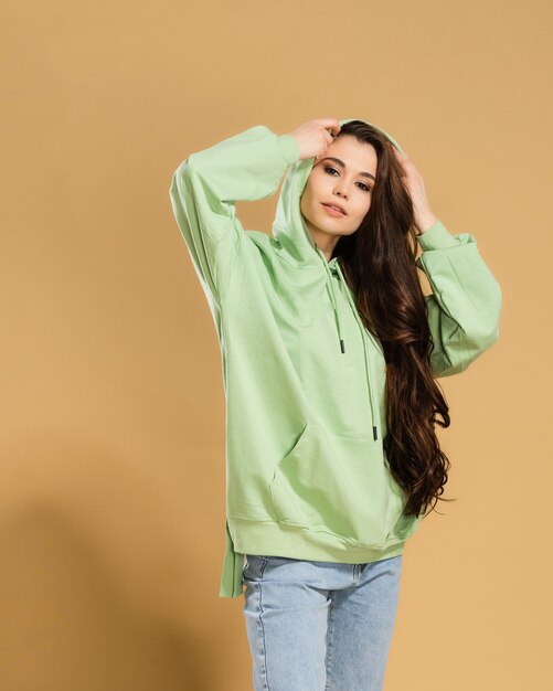 Young beautiful girl with long hair in a pistachio-colored hoodie straightens the hood on a pastel orange.