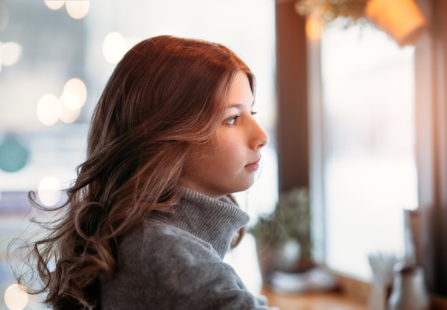 A young beautiful girl with long hair sits at a table in a cafe and looks out the window