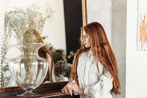 La giovane bella ragazza con capelli lunghi sta vicino allo specchio di mattina nella camera da letto