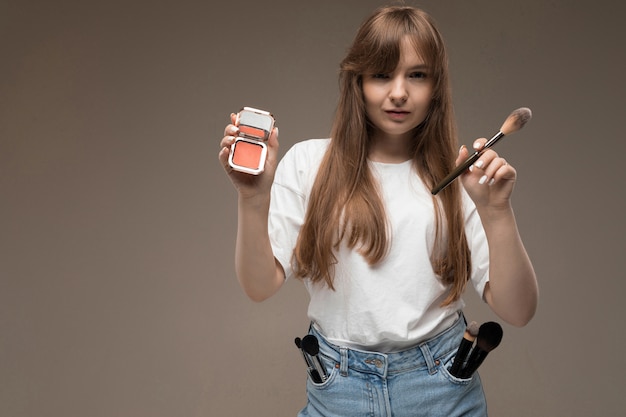 A young beautiful girl with long wavy rustic hair, nud makeup, in a white jersey, holds a makeup brush and blush, and a lot of makeup brushes in her pockets gin